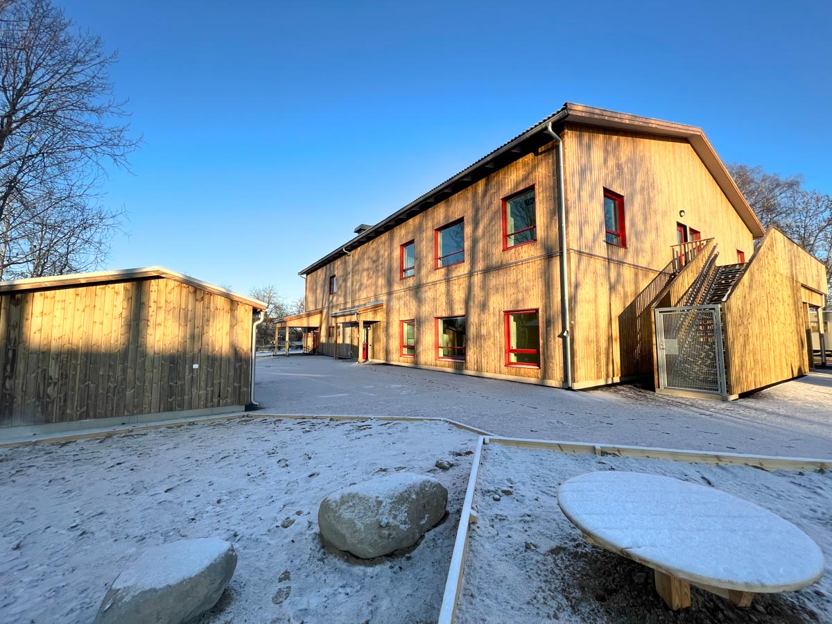 Förskolan snett från sidan. Byggnaden är i trä och består av två våningar. I förgrunden syns en sandlåda som är täckt av snö. 