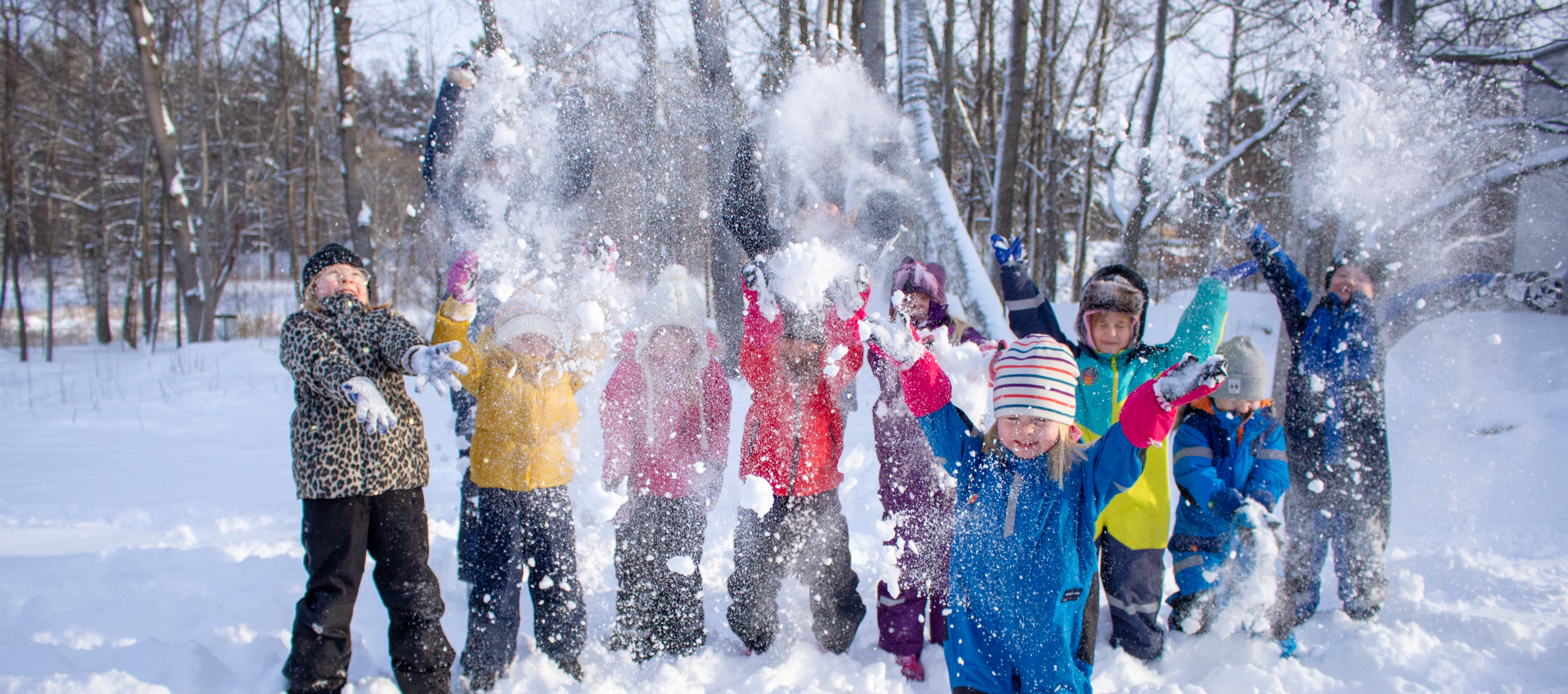 Barn som står utomhus i färgglada overaller och kastar snö. 